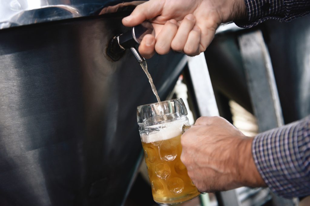Strong man's hands pour beer in tumbler from beer tap at craft brewery. Brewer pours beer into glass for trial of consignment.