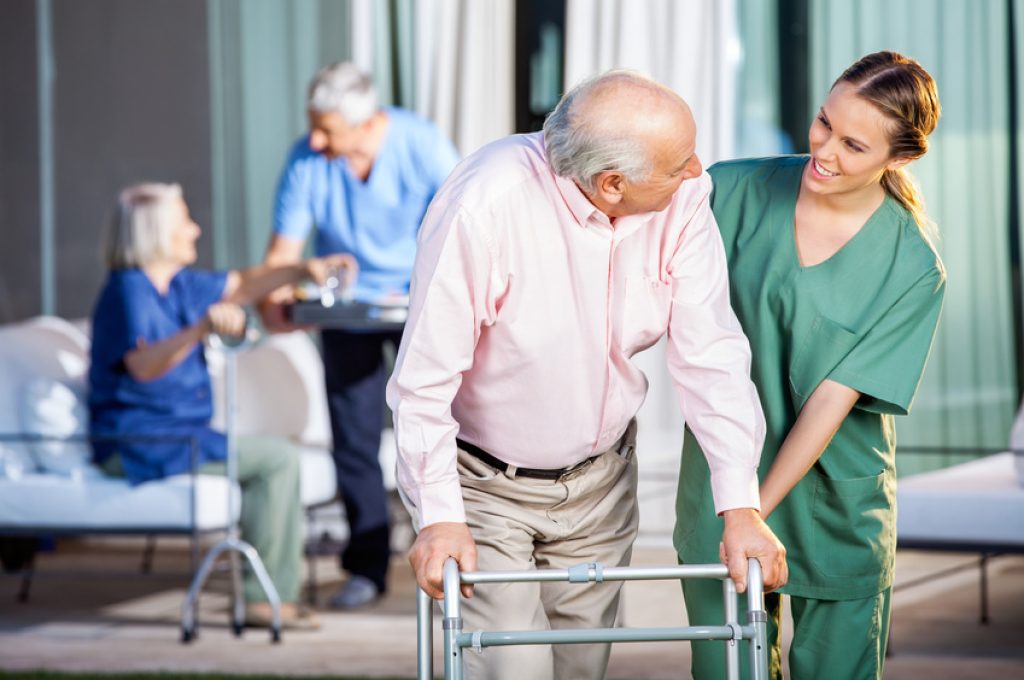 Happy female caretaker assisting senior man in using Zimmer frame at nursing home yard