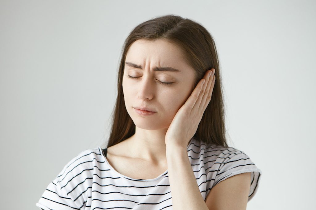 Studio shot of attractive casually dressed young dark haired female keeping eyes closed and covering sore ear with hand while having some problems with hearing. Health, people and pain concept